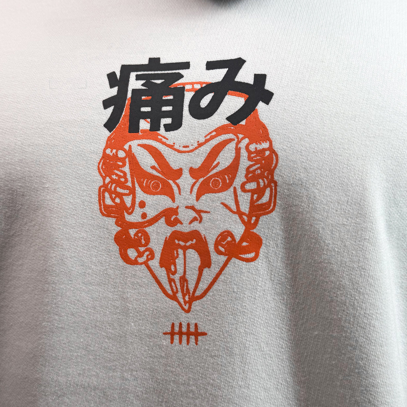 A close-up of a white crewneck sweatshirt featuring a graphic print of a red and black mask with Japanese characters, shown on a model wearing a black respirator mask.